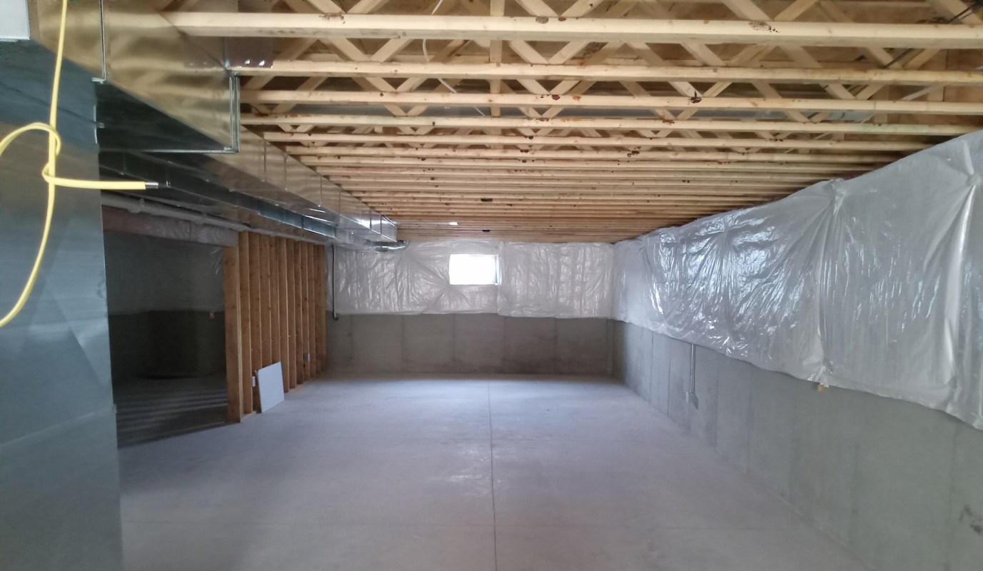 An unfinished basement with exposed beams, insulation, and plastic sheeting along the walls.