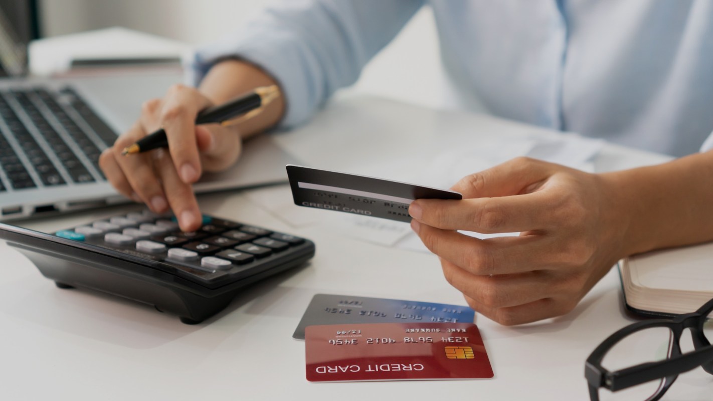 A person holds a credit card while using a calculator and laptop on a desk with papers and glasses nearby.