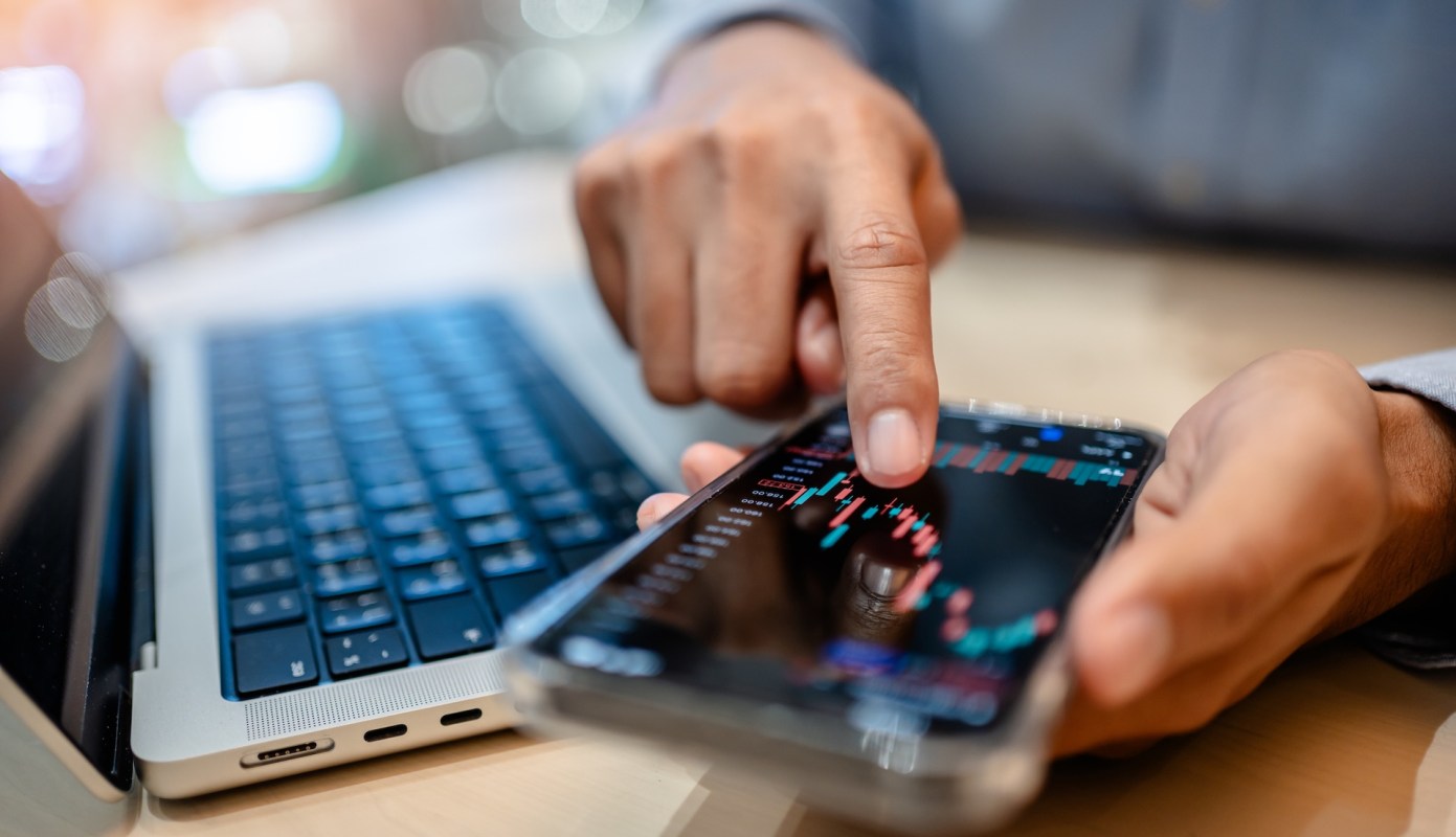 A person points at a smartphone displaying financial data, with a laptop in the background.
