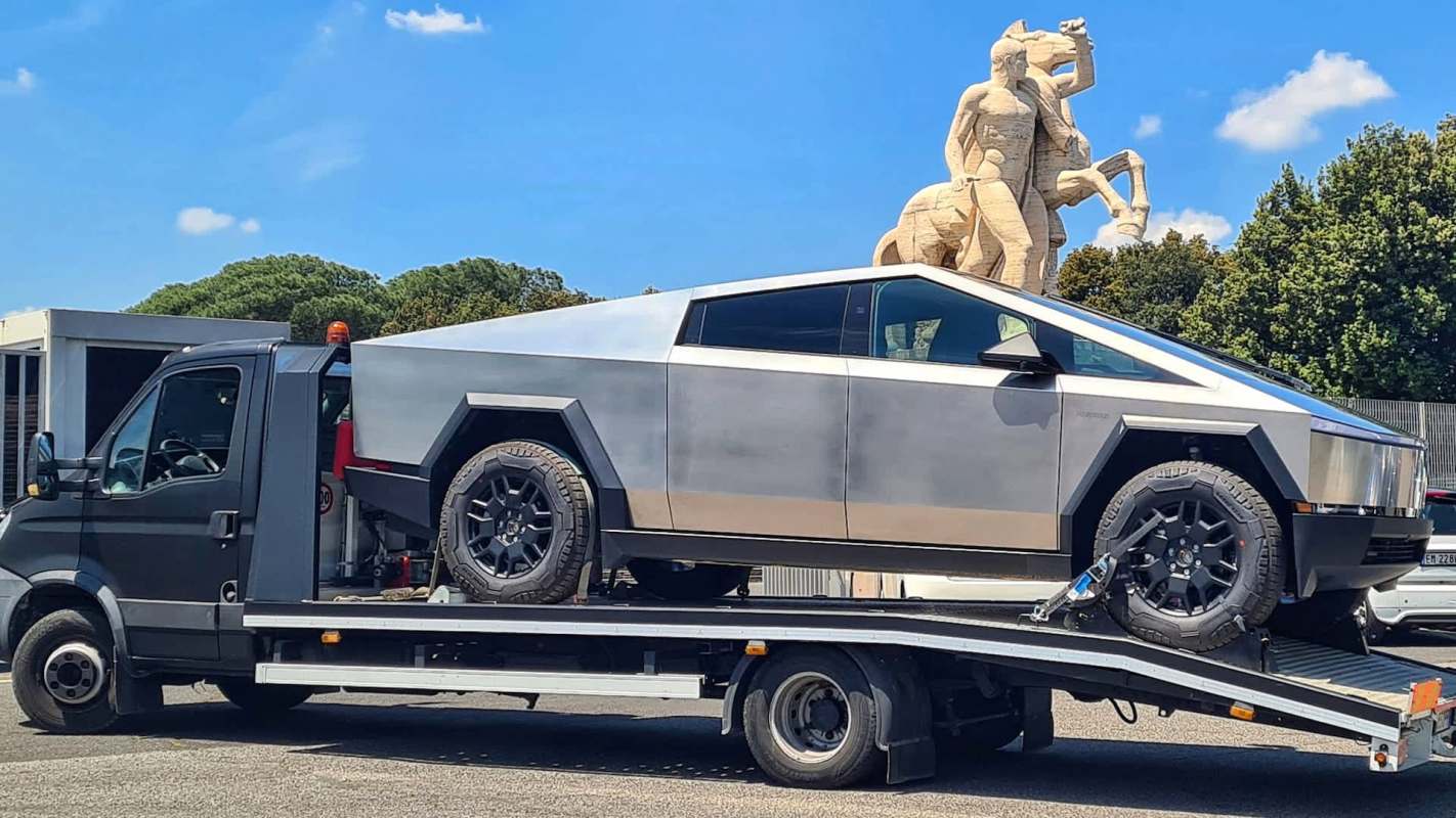 A Tesla Cybertruck on a tow truck with a sculpture in the background.