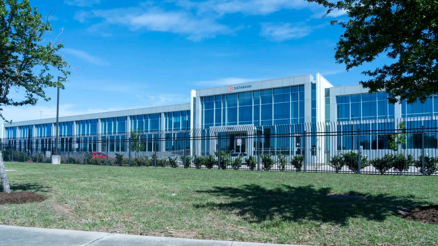 A data center building with large glass windows and a gated entrance, surrounded by grass and trees.