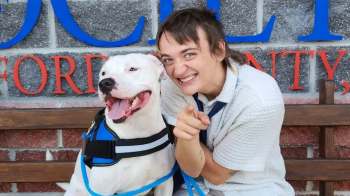 A person and a dog sit on a bench in front of a brick wall with a sign.