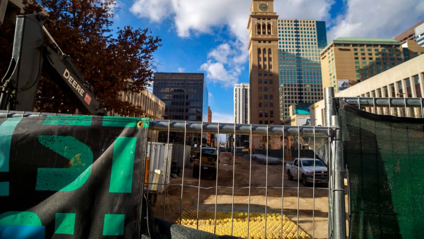 A construction site in Denver with a fence, heavy machinery, and tall buildings in the background under a partly cloudy sky.