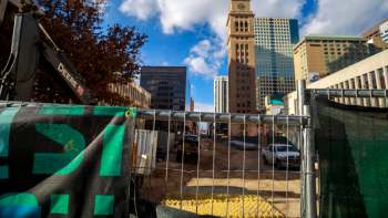 A construction site in Denver with a fence, heavy machinery, and tall buildings in the background under a partly cloudy sky.