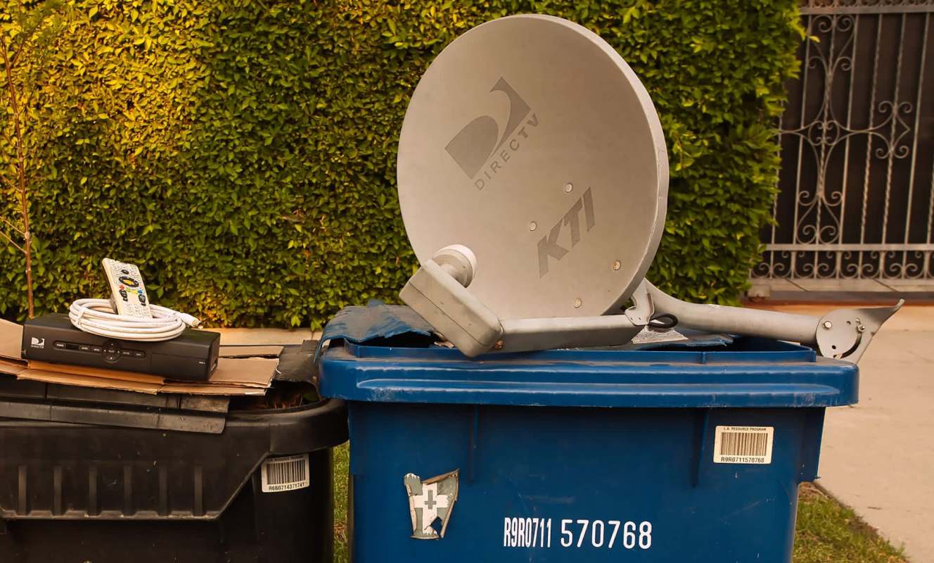 A DirecTV satellite dish and a cable box sit on top of a blue trash can, alongside a remote control.