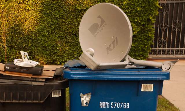 A DirecTV satellite dish and a cable box sit on top of a blue trash can, alongside a remote control.