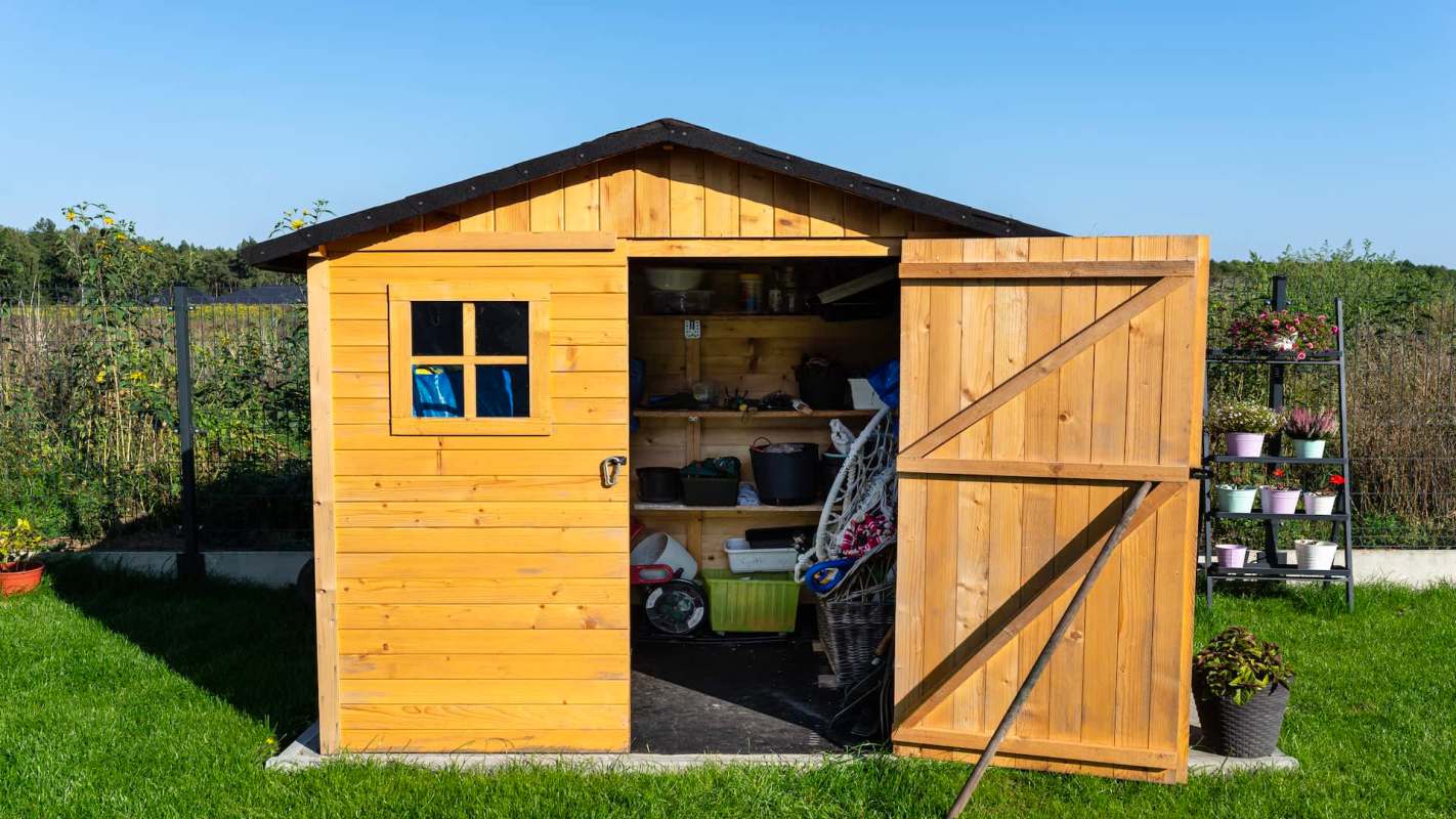A wooden shed with an open door showing tools and storage, beside a tiered planter with flowers.