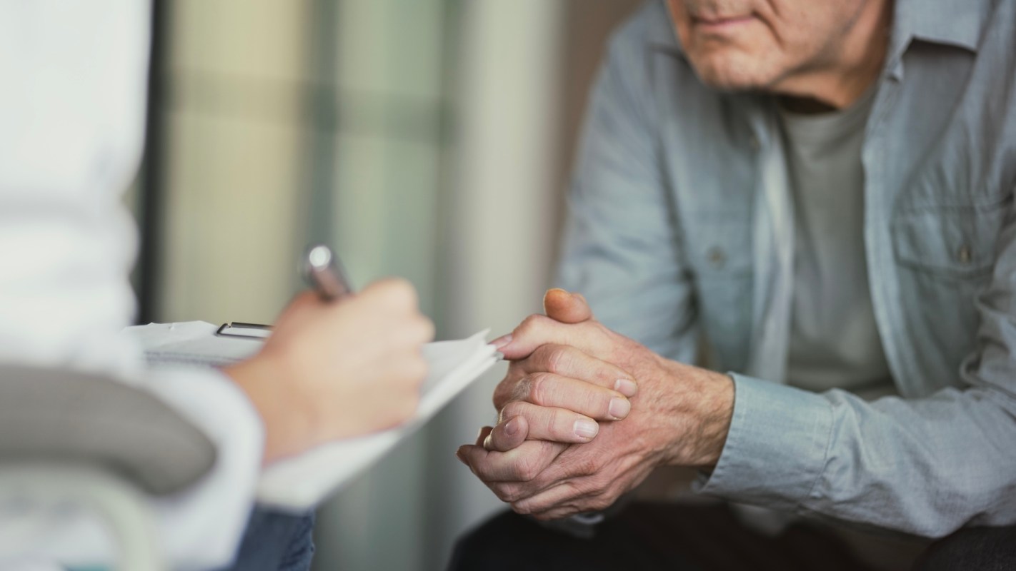 A person writes on a notepad while another person rests their hands together.