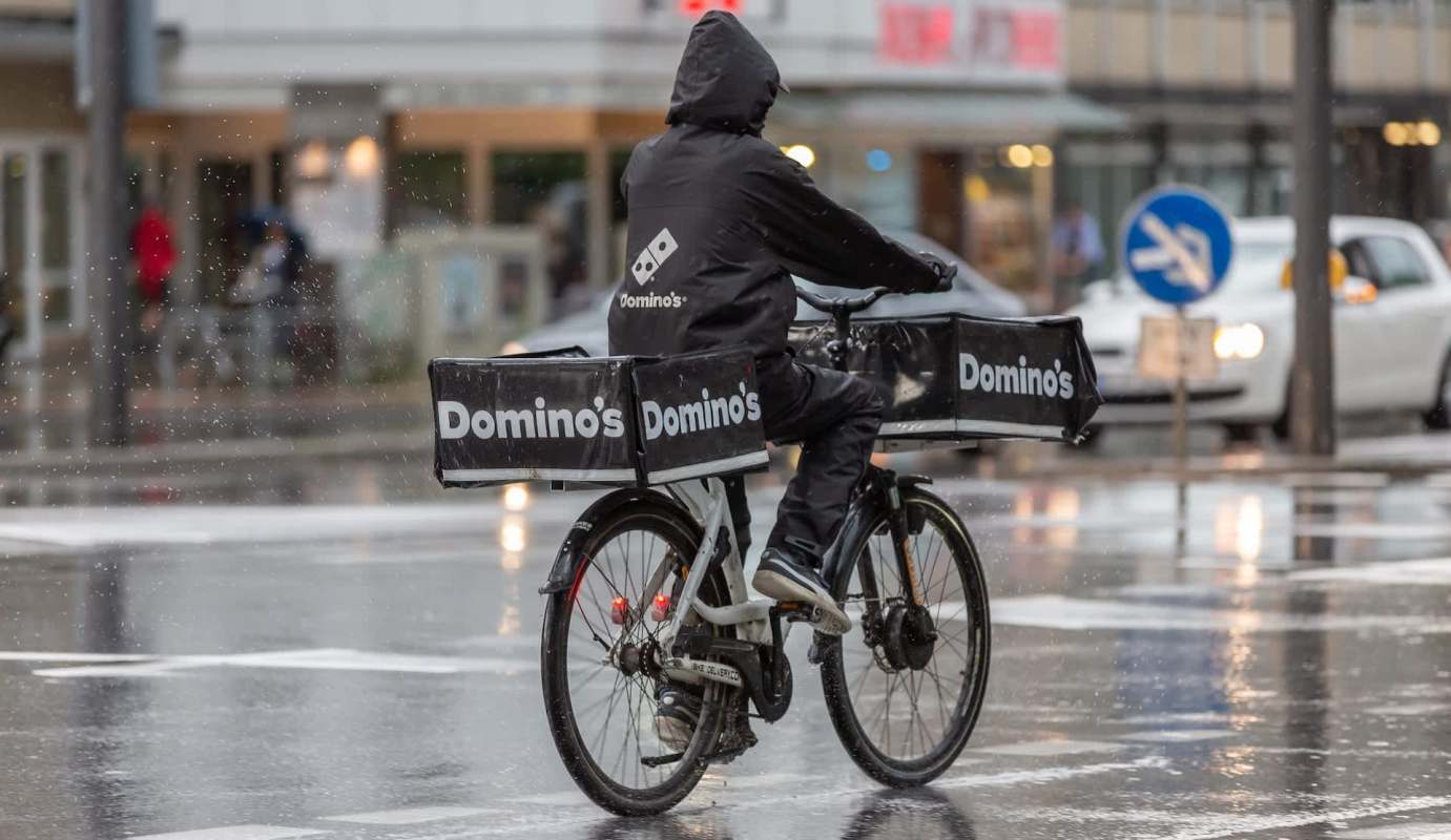 A delivery person on a bicycle rides through rain, carrying Domino's pizza boxes.