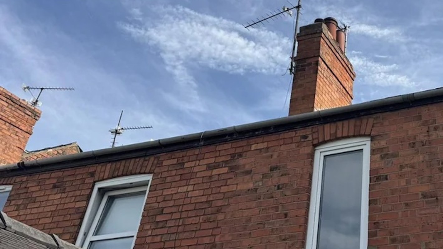 A brick wall with windows, a chimney, and rooftops under a blue sky with clouds.