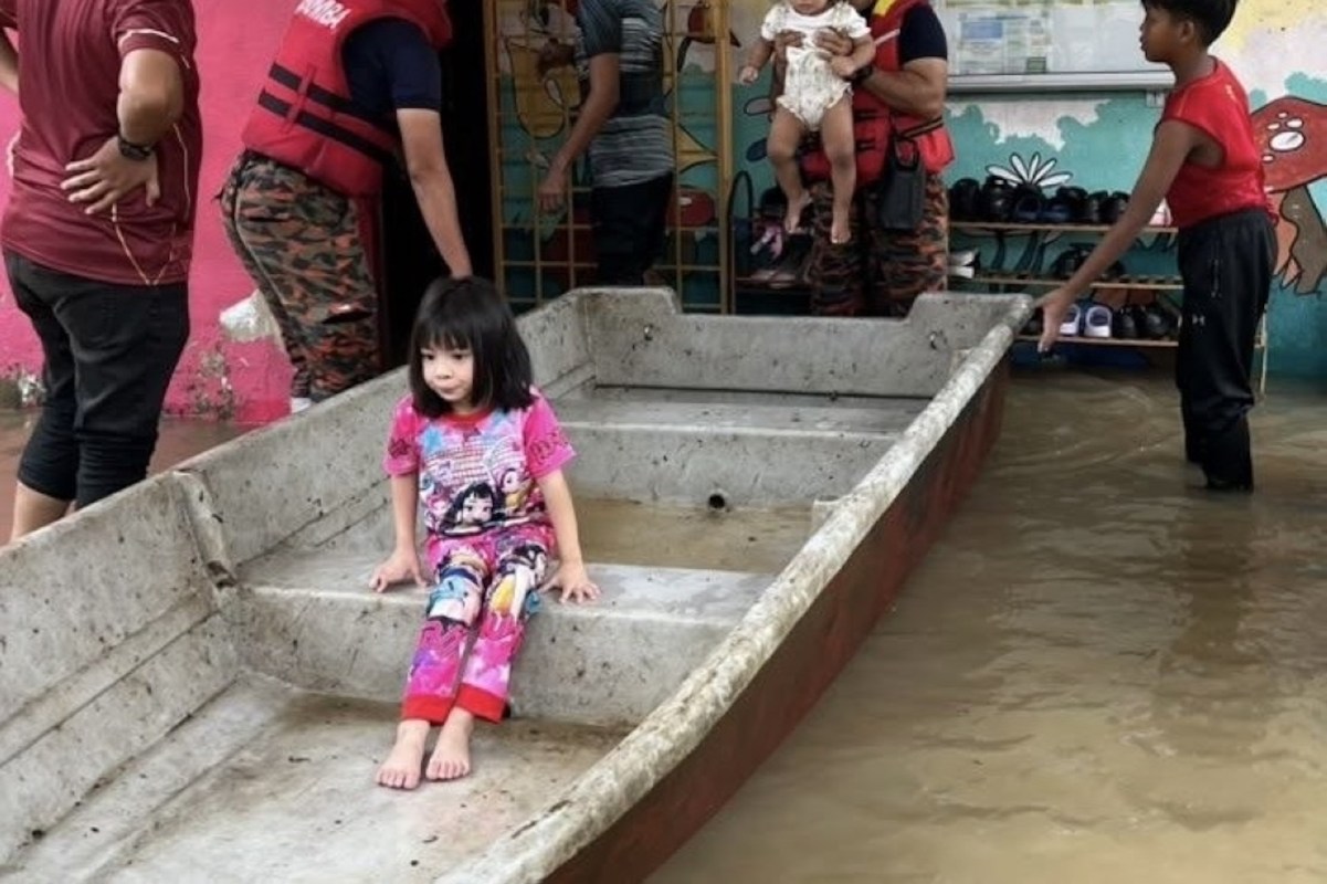 A girl sits in a boat inside a flooded area, while adults assist others nearby.
