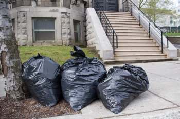 Three large black trash bags are placed next to a tree on a sidewalk near a set of stairs.