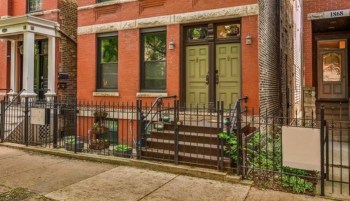 A brick building entrance with green doors and a fenced front yard.