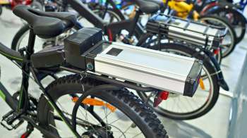 A bicycle with a battery mounted on the rear rack in a shop with other bicycles in the background.