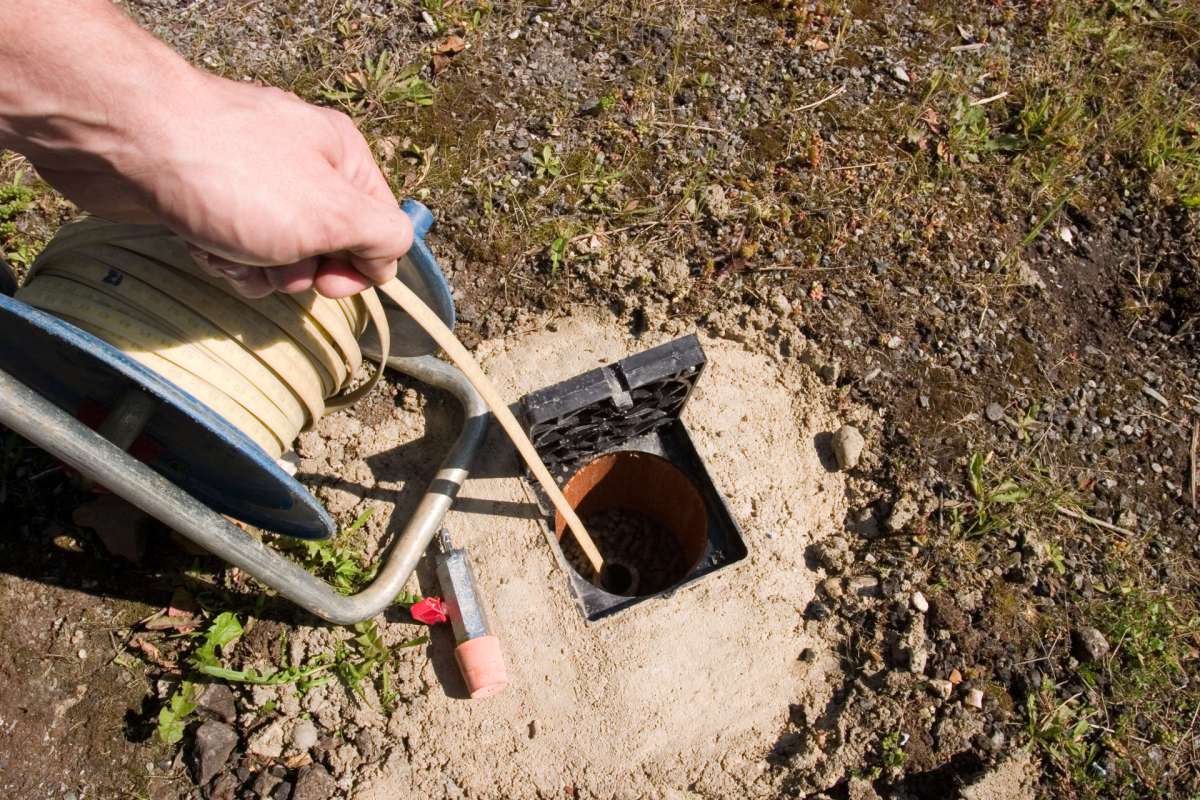 A hand uses a tool to work near an open access point in the ground next to a reel of tape.