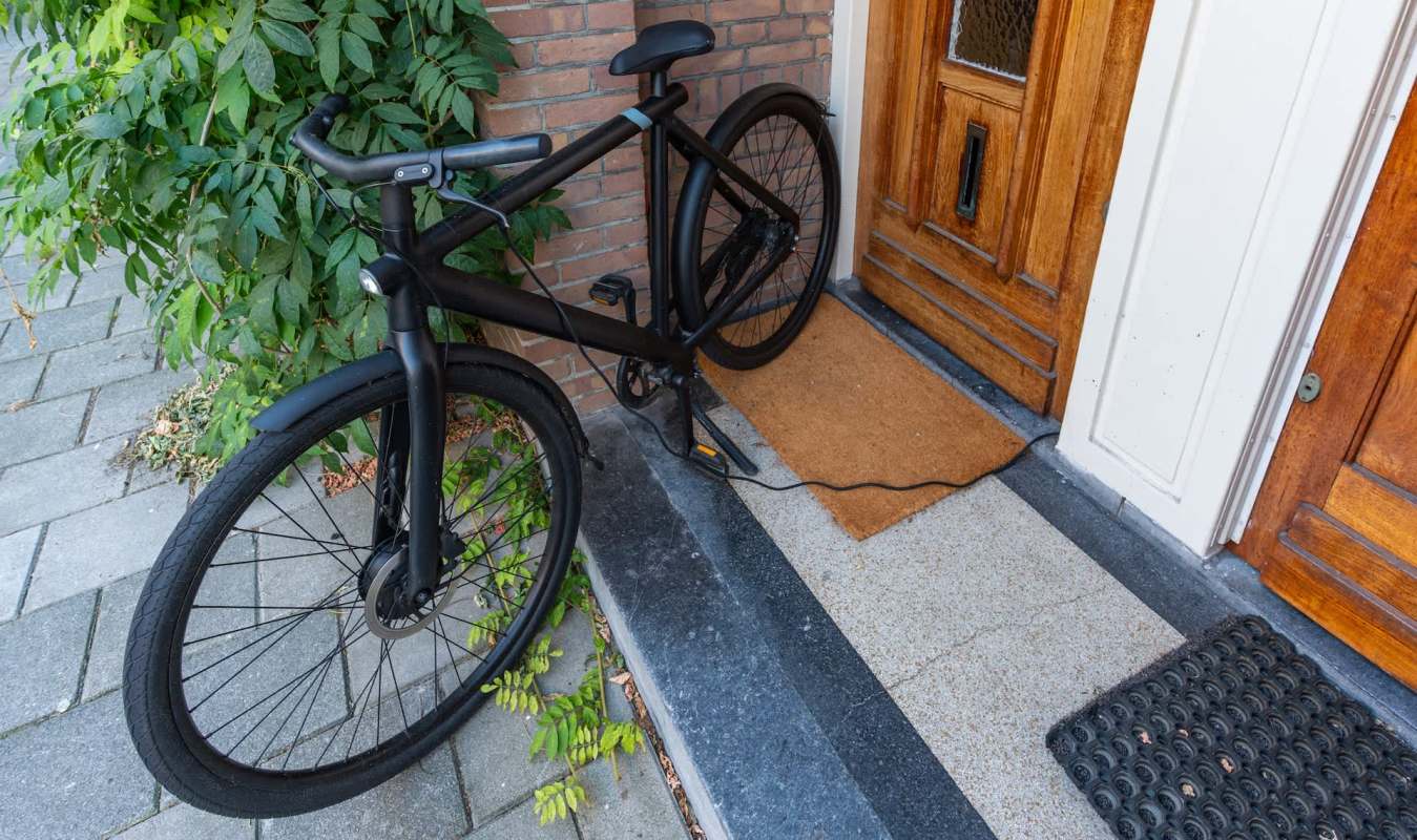 A black electric bicycle charging in front of a door with a doormat and surrounding greenery, with a cord under the door.