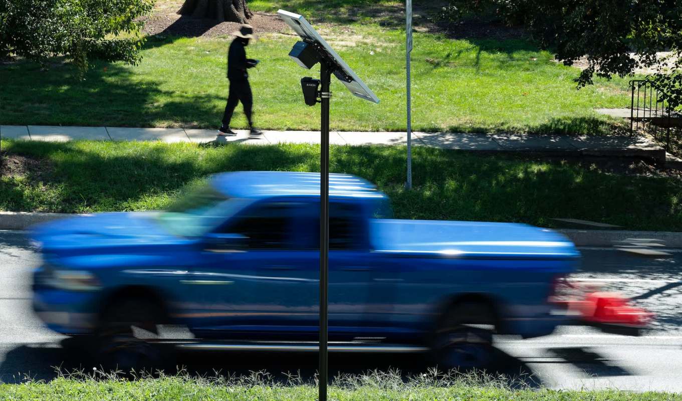 A blue pickup truck drives past a Flock Safety camera with a solar panel while a person walks on the sidewalk.
