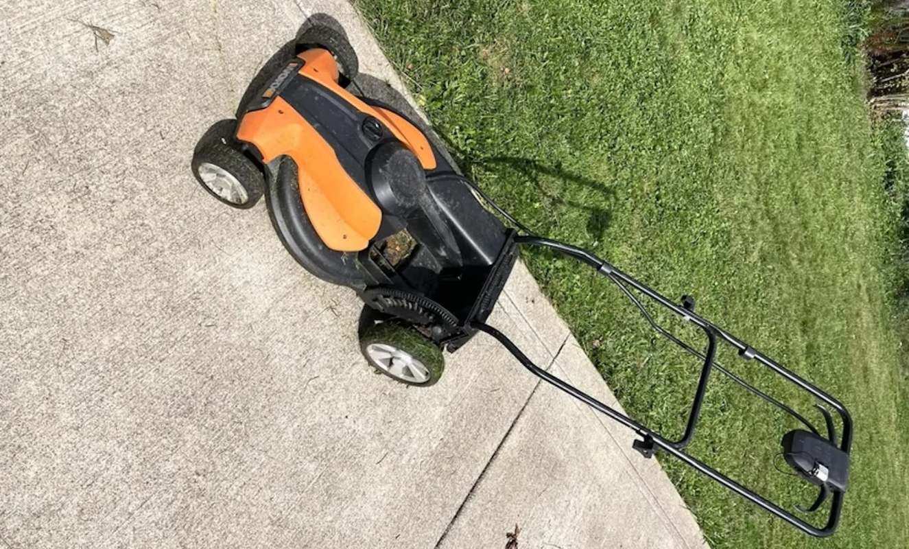 An orange and black electric lawn mower on a concrete surface next to green grass.