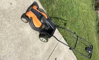 An orange and black electric lawn mower on a concrete surface next to green grass.