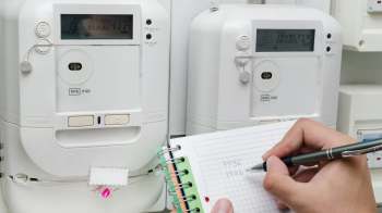 A hand writes numbers in a notebook next to two electric meters on a wall.