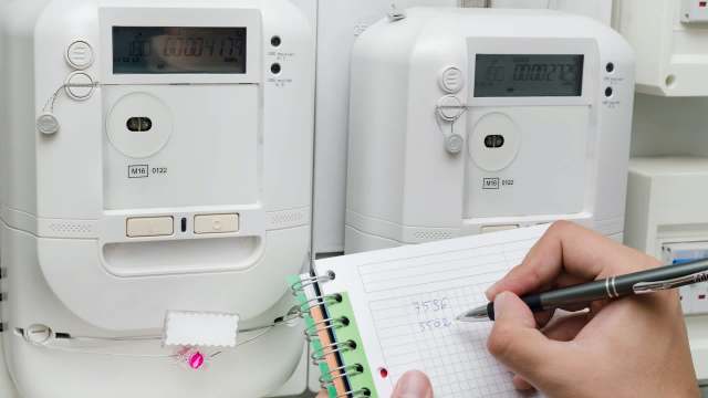 A hand writes numbers in a notebook next to two electric meters on a wall.