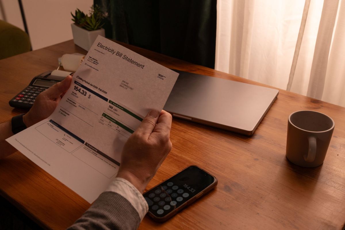 A person holding an electricity bill statement with a calculator and laptop on a wooden table.