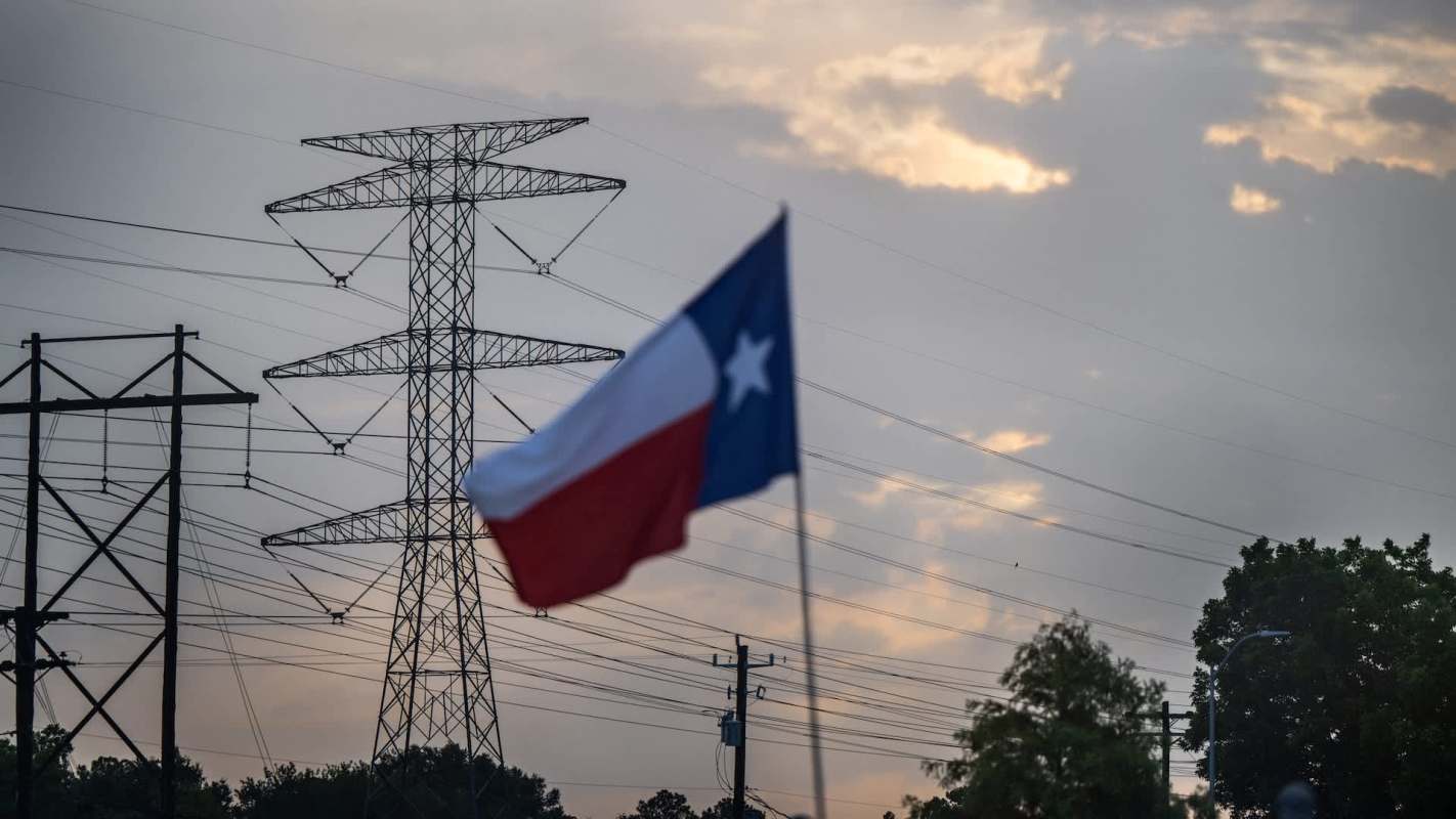 A Texas flag in the foreground with power lines and a cloudy sky in the background.