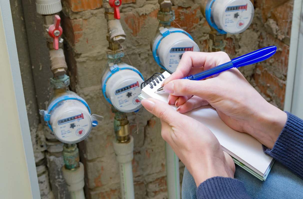 A person writes in a notebook while recording information from water meters on a wall.