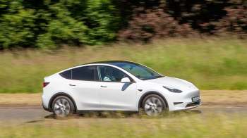 A white electric car drives along a rural road surrounded by tall grass and trees.