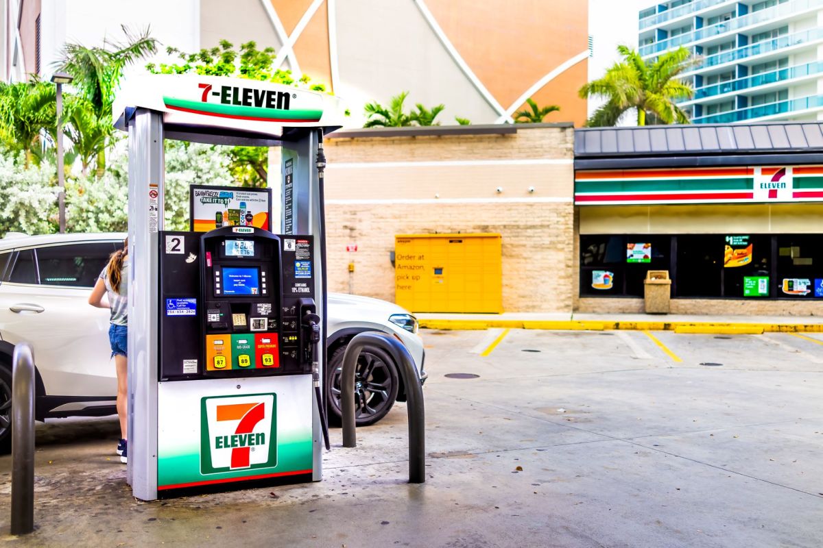 A 7-Eleven gas pump next to a white car with a convenience store in the background.
