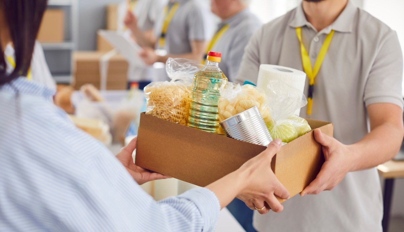 A person hands a box filled with food items to another person in a community setting.