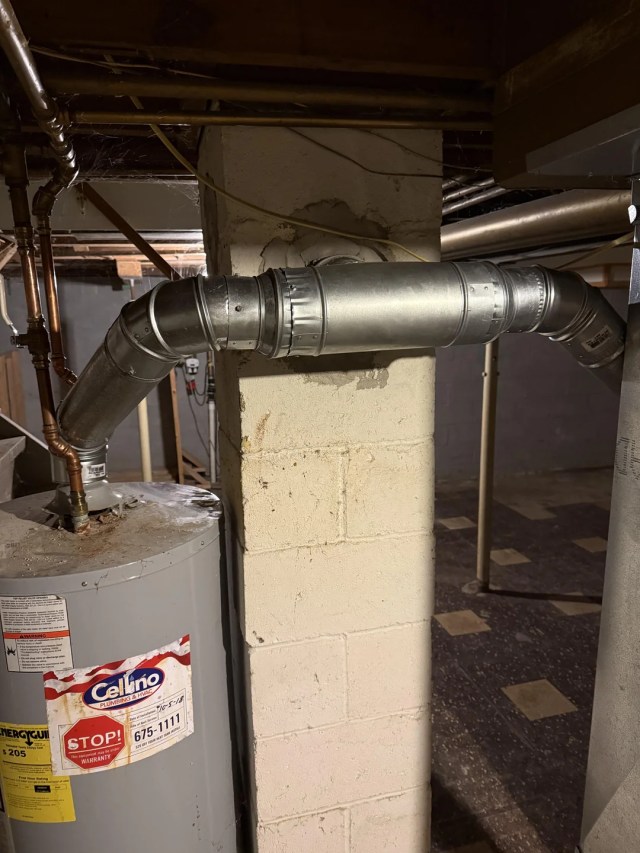 A metallic ventilation duct connected to a wall in a basement setting.