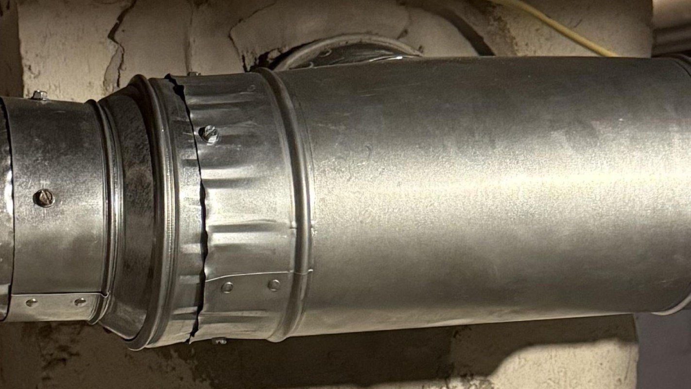 A metallic ventilation duct connected to a wall in a basement setting.