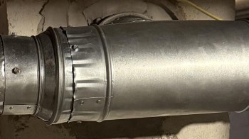 A metallic ventilation duct connected to a wall in a basement setting.
