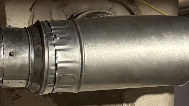 A metallic ventilation duct connected to a wall in a basement setting.