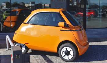 An orange compact electric car parked near a glass building reflecting other vehicles.