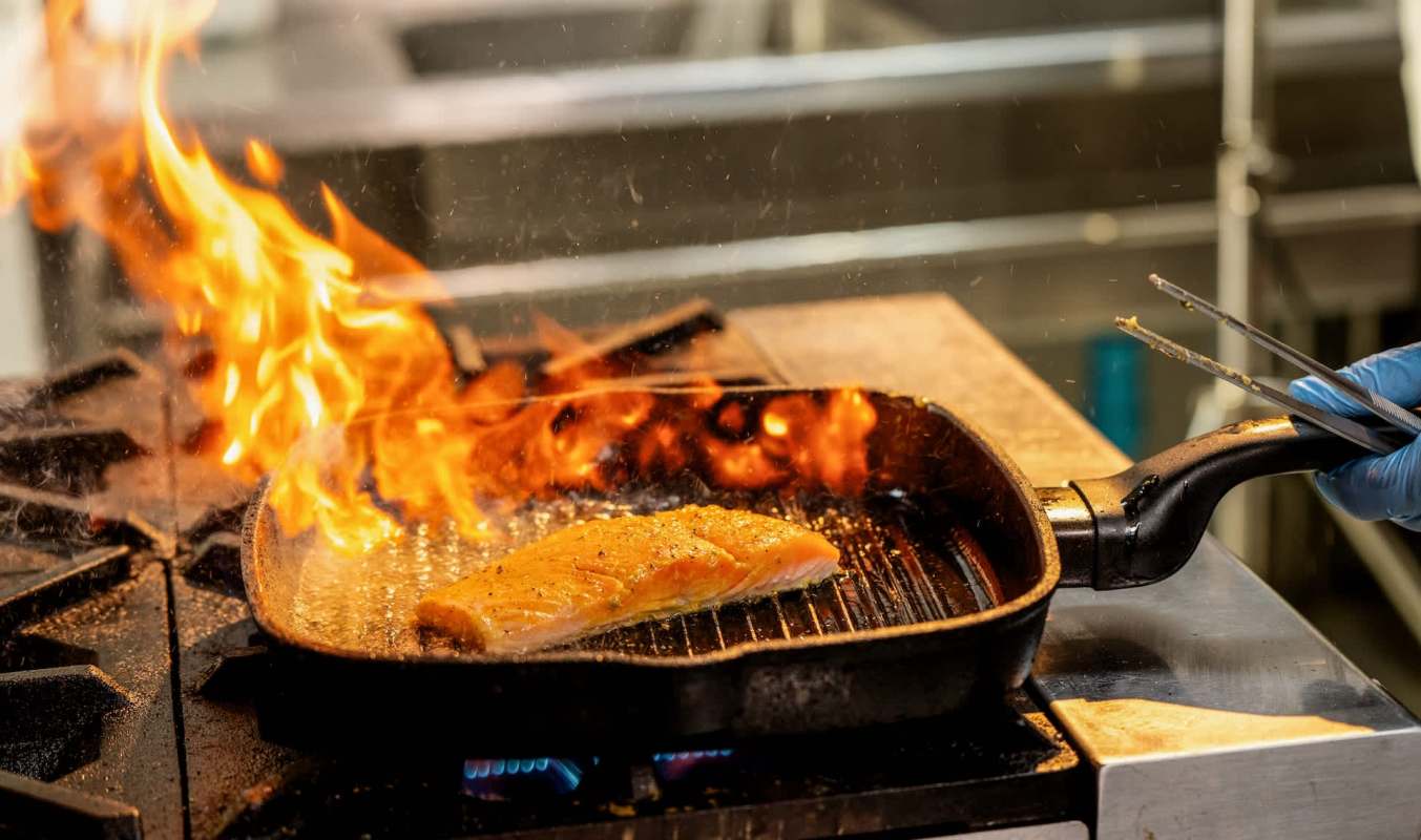 A piece of salmon cooks in a sizzling pan with blue gas flames and oil spatters.