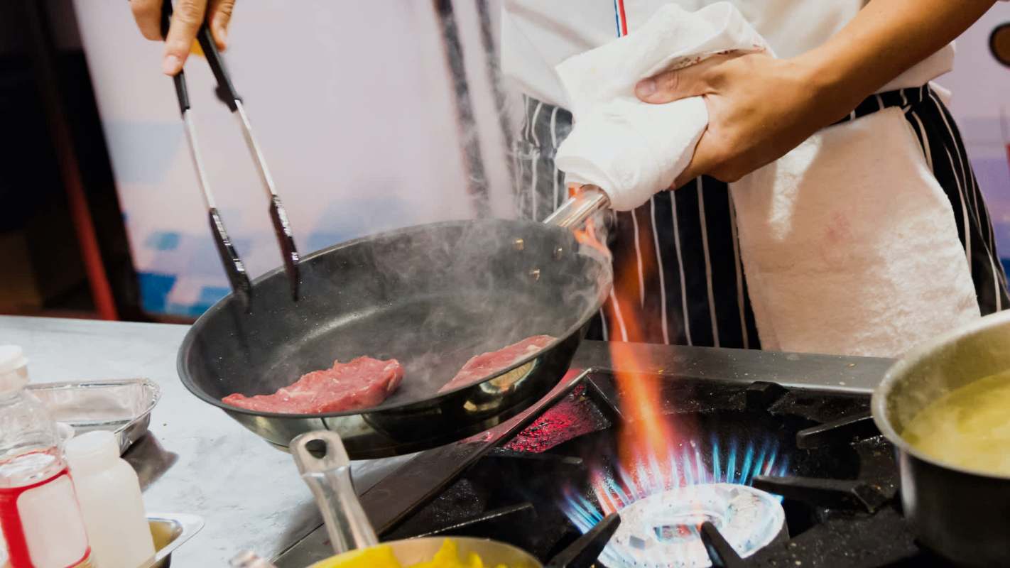 A chef uses tongs to sauté meat in a pan over a gas stove with visible flames.