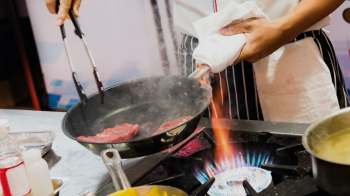 A chef uses tongs to sauté meat in a pan over a gas stove with visible flames.