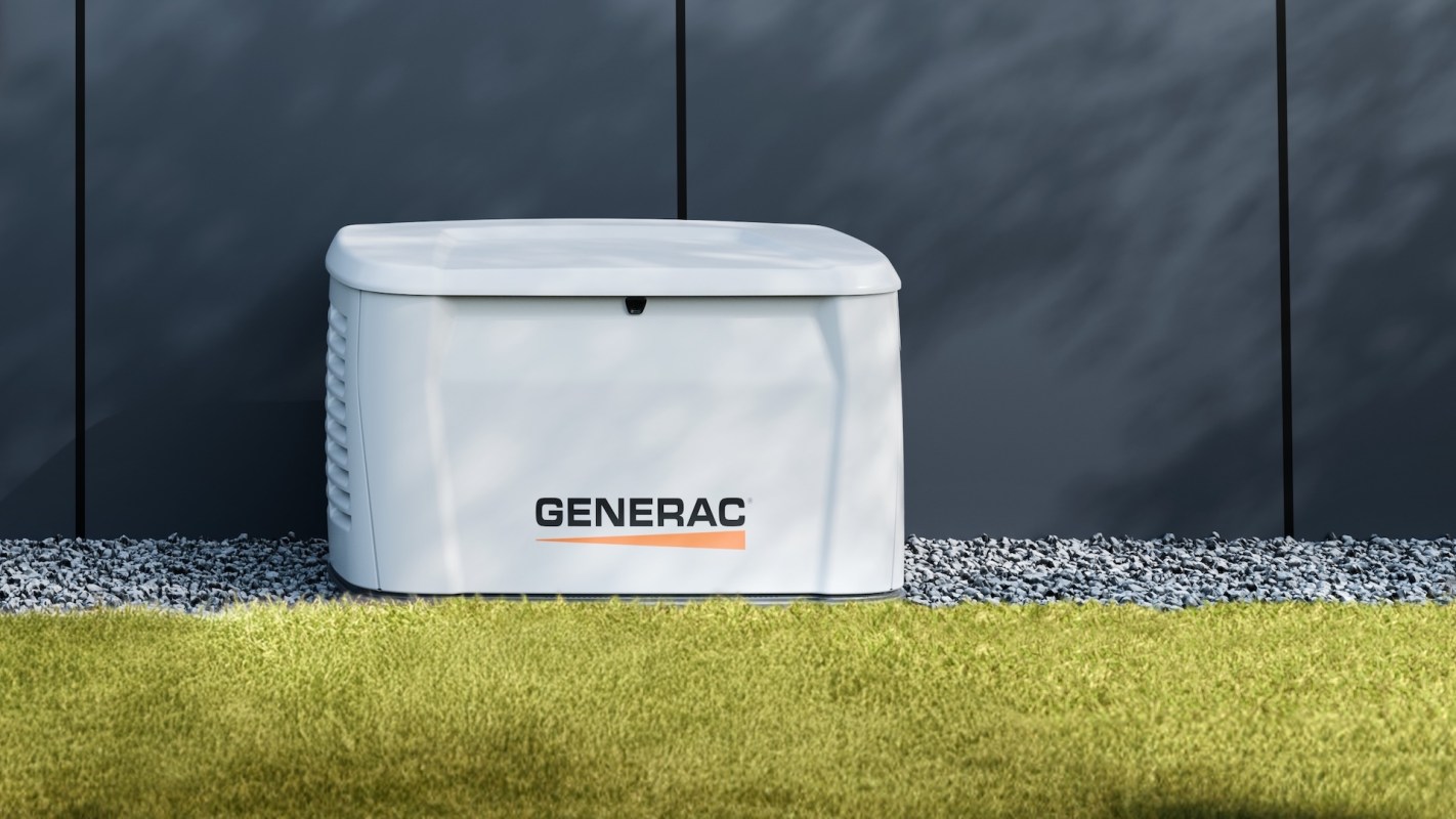 A Generac generator unit on a gravel surface beside a grass area and a gray wall.