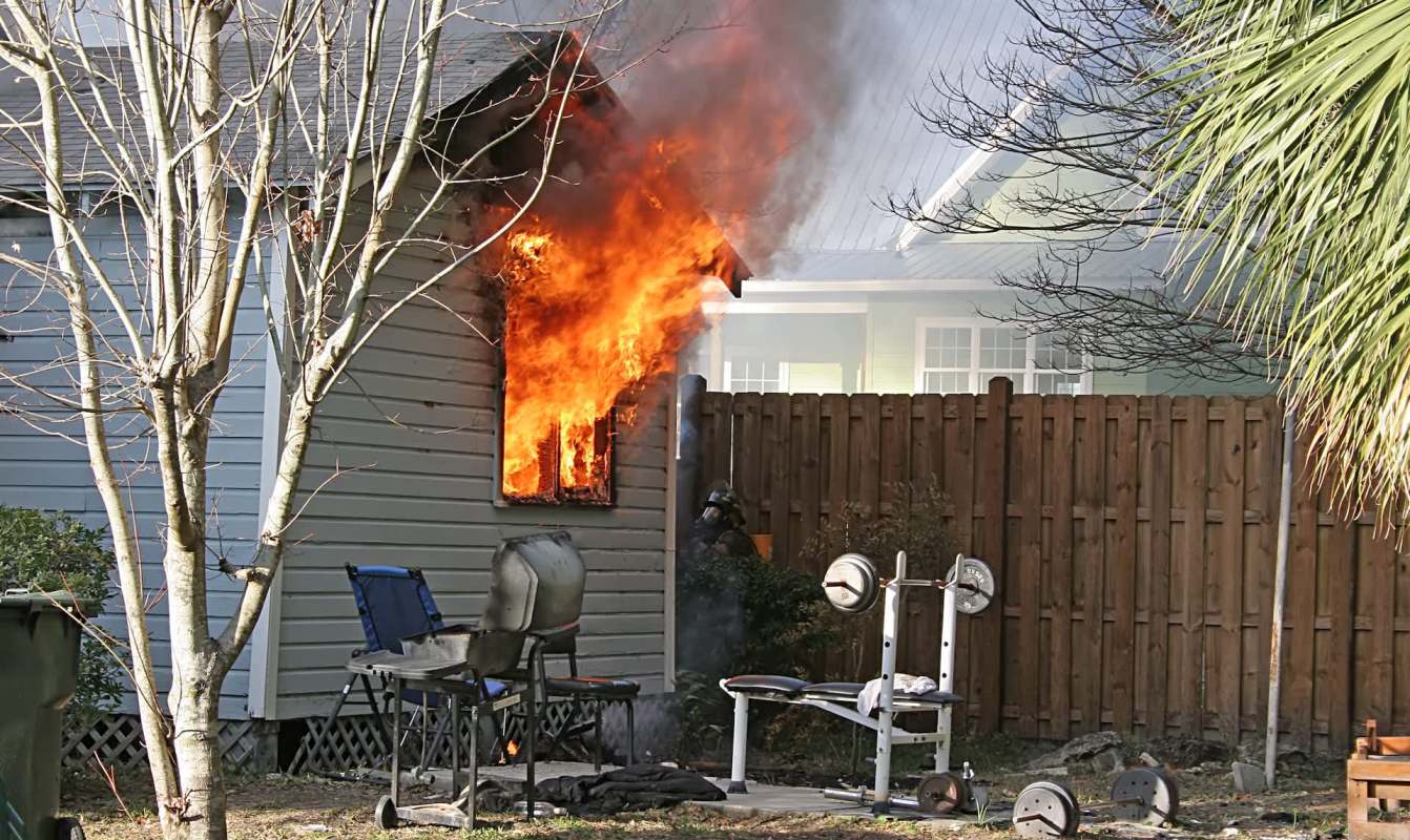 A house is on fire at a window, with outdoor furniture and workout equipment in the foreground.