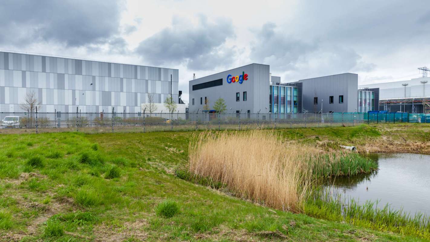 A Google building beside a pond with grass and reeds in the foreground under a cloudy sky.