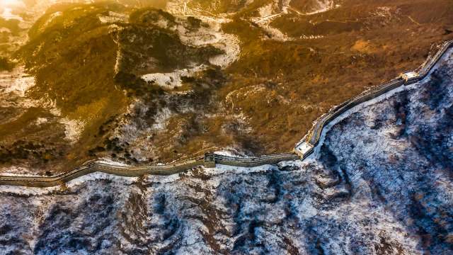 The Great Wall of China extends across rocky terrain with patches of snow and vegetation.