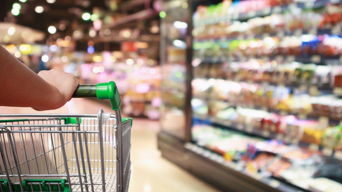 A hand grips a shopping cart in a grocery store aisle filled with colorful products.
