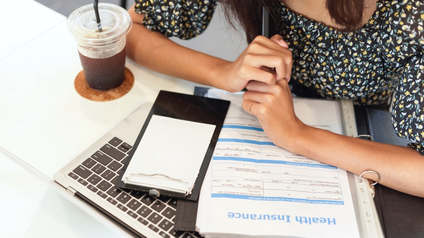 A person sits at a table with a laptop, clipboard, and a drink, reviewing health insurance documents.
