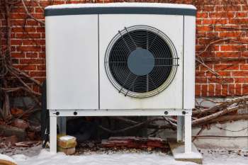 An outdoor heat pump unit placed against a brick wall with exposed vines and snow on the ground.
