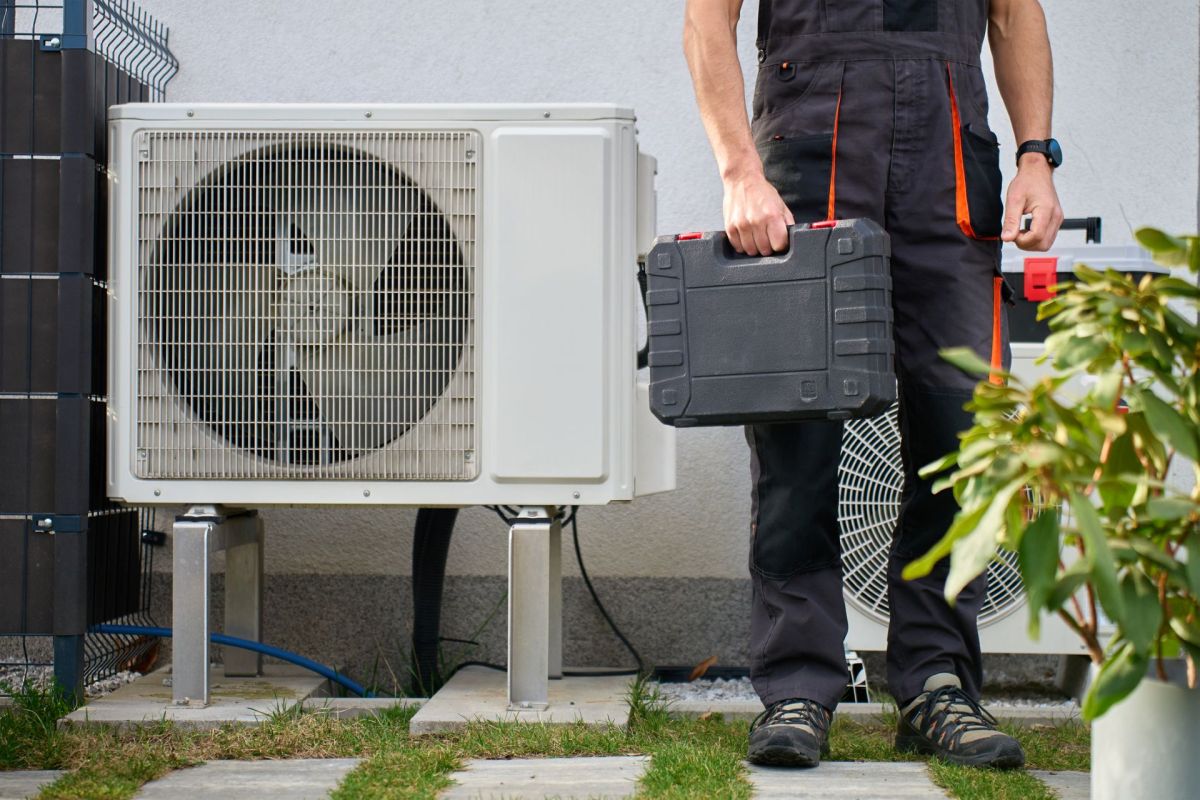 A technician stands beside an air conditioning unit while holding a tool case.