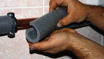 A person installs foam insulation onto a copper pipe mounted on a wall.