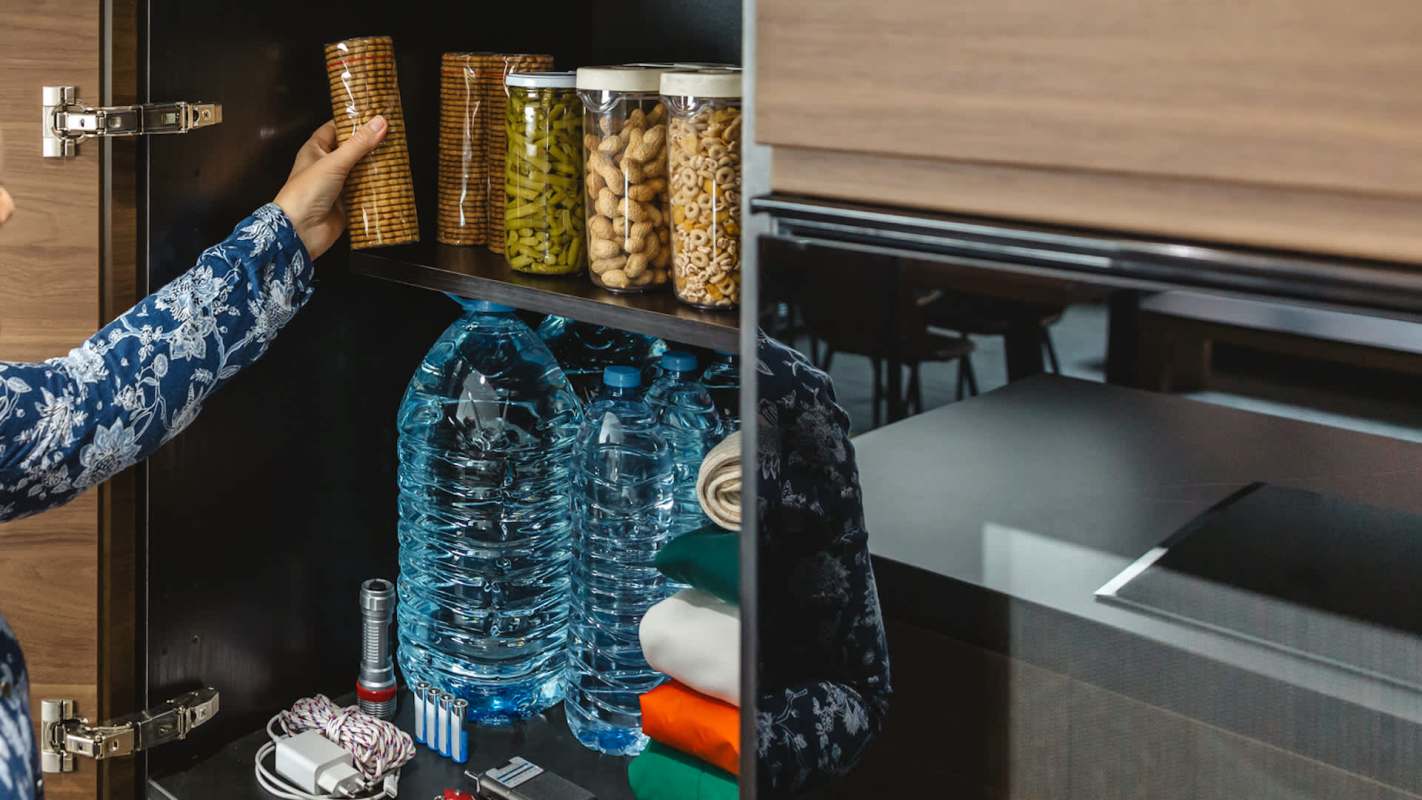 A person reaches for jars of food and snacks in a kitchen cabinet with water bottles and various items on the shelf.