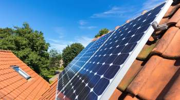 A solar panel installed on a tiled roof with trees and a blue sky in the background.